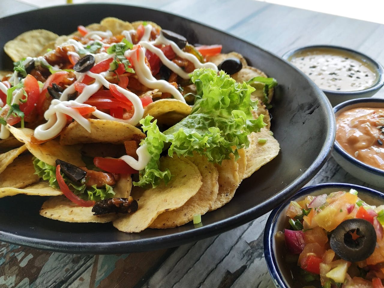 corn chips, green lettuce, sliced black olives,, tomatoes & red bell pepper coverd in mayonnaise on a black plate with some sauces on the side